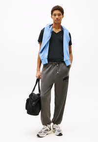 Schwarzes V-Ausschnitt-T-Shirt, hellblauer Pullover über die Schultern gelegt, graue Jogginghose mit "TOMMY"-Logo, schwarze Tasche und graue Sneaker mit weißen Akzenten.