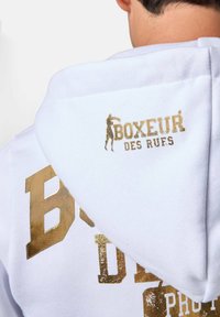 Felpa bianca con testo e grafica dorata sul retro, con la scritta "BOXEUR DES RUES". Tessuto morbido con finitura testurizzata e cappuccio regolabile.