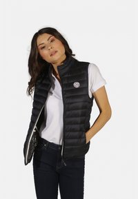 Doudoune noire à zip sur le devant, design matelassé et patch logo, portée sur une chemise blanche et un jean bleu. Texture lisse et coupe ajustée.