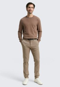 Homme se tenant avec les mains dans les poches, portant un pull marron, un pantalon beige, une ceinture noire et des baskets blanches sur un fond clair.