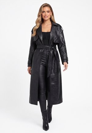 Kvinde med langt blondt hår iført en sort faux-læder trenchcoat med bælte, sort top, sorte højttaljede bukser og sorte ankelstøvler.