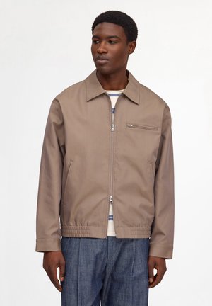 Junger Mann trägt eine beige Jacke mit Reißverschluss über einem gestreiften Hemd und blauen Hosen, steht vor einem einfarbigen weißen Hintergrund.