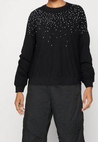 Sudadera negra con brillantes dispersos en la parte frontal superior, mangas anchas y puños y dobladillo acanalados. Textura de tela suave.