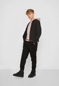 Veste bomber noire sur un hoodie rose clair, associée à un pantalon cargo noir et des baskets montantes noires, sur fond blanc.