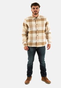 Geruite shirt in beige en wit, lange mouwen, met knopen aan de voorkant, enkele zak. Gecombineerd met donkere jeans en bruine veterschoenen.