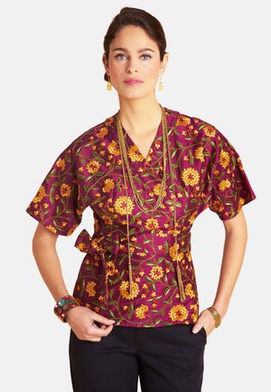 Woman wearing a purple wrap-style top with yellow floral embroidery, layered gold necklaces, and colorful bracelets, posing with hands in pockets.