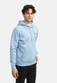 Sudadera azul claro hecha de una suave mezcla de algodón, con un bolsillo tipo canguro en la parte frontal y un pequeño logo rojo en el pecho. Diseño de corte relajado.