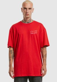 Červené bavlněné oversized tričko s krátkým rukávem, na levé straně s bílým nápisem "ZAVOLEJTE MÉMU PRÁVNÍKOVI". Tkanina vypadá hladce.