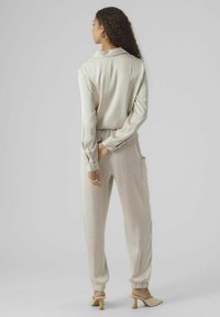 Ensemble de loungewear beige composé d'un haut à manches longues et d'un pantalon fuselé. Texture lisse, détails de col et poches latérales. Des mules à talons complètent le look.