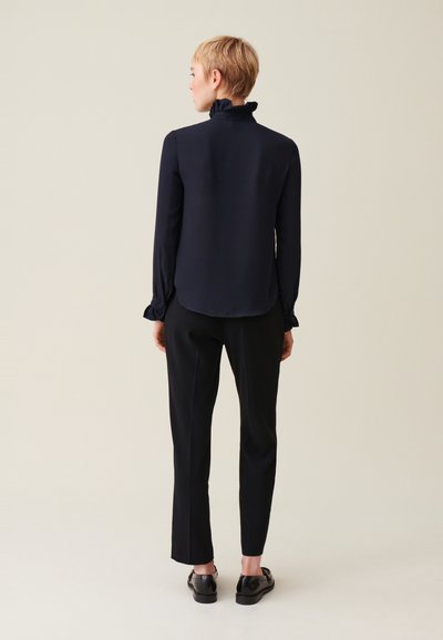 Blusa navy a maniche lunghe con dettagli di colletto arricciato, abbinata a pantaloni neri e scarpe nere, vista da dietro su uno sfondo neutro.