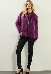 Blusa viola trasparente con maniche lunghe e fiocco al collo, abbinata a jeans skinny neri e tacchi neri. Finiture in tessuto liscio e lucido.