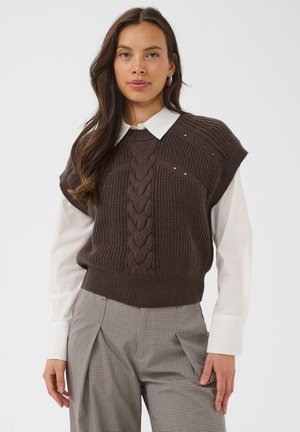 Femme portant un gilet torsadé marron sur une chemise blanche à col et un pantalon à carreaux gris, debout devant un fond uni clair.