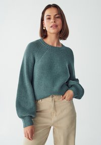 Tiefgrüner gestrickter Pullover mit Rippendesign, Rundhalsausschnitt und Puffärmeln, kombiniert mit beigen hoch geschnittenen Hosen.