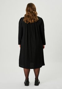 Schwarzes plissiertes Kleid mit langen Ärmeln, gerafftem Rücken und lockerer Silhouette. Getragen mit schwarzen Schuhen und Strumpfhosen.