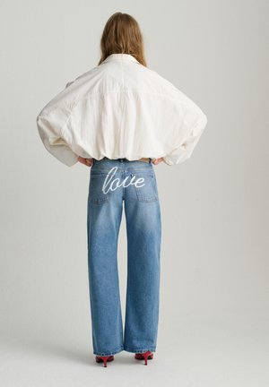 Personne aux cheveux longs vue de dos, portant une chemise blanche surdimensionnée, un jean bleu avec le mot « love » brodé au dos, et des chaussures à talons rouges.