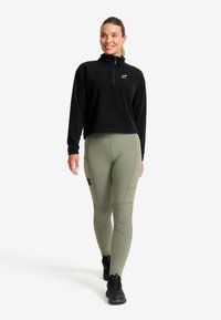 Svart zip-up fleece-pullover med hög krage, kombinerad med ljus olivgröna åtsittande leggings med texturerade paneldetaljer. Svarta sneakers.