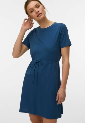 Woman adjusting hair wearing a short-sleeve blue dress with waist tie against a plain white background.