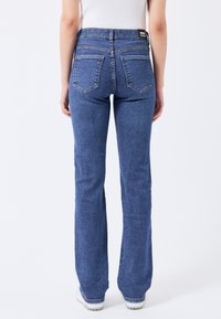 Jeans de mezclilla acampanados en azul oscuro, con cintura alta, bolsillos traseros tradicionales y una textura suave. Etiqueta visible en la cinturilla.