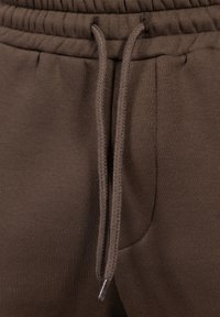 Hnědé joggingové kalhoty s elastickým pasem, vyrobené z měkké látky, s kapsou na boku a hladkým povrchem.