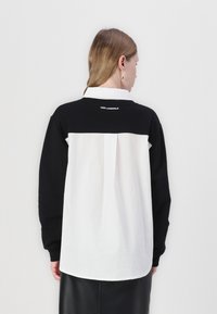 Svart och vit skjorta med långa ärmar; svart övre del, vit nedre del, synliga sömdetaljer och logotyp på baksidan.