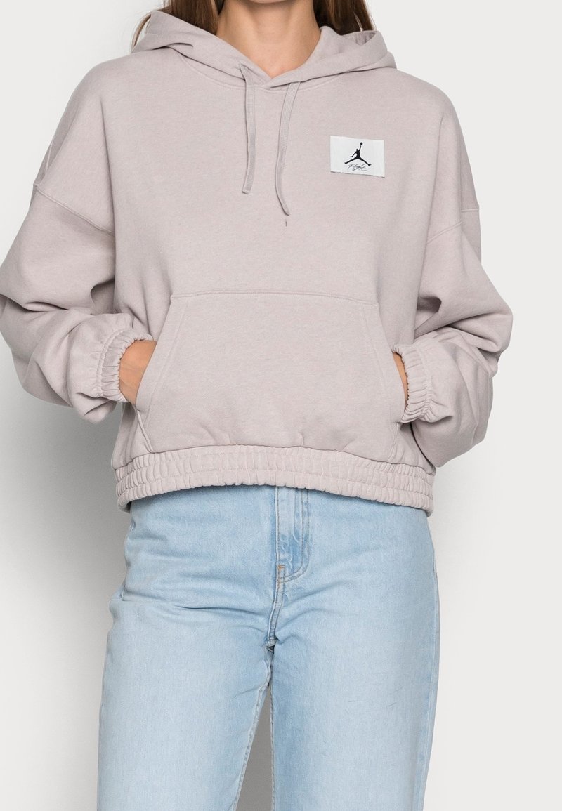 Femme portant un sweat à capuche beige clair avec poche avant et patch logo Jordan, associée à un jean bleu clair, mains dans les poches.
