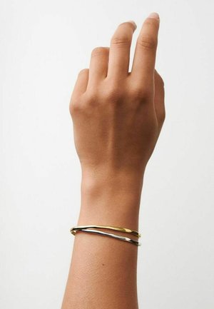 Main avec les doigts légèrement pliés, portant deux bracelets métalliques fins de forme irrégulière, un en or et un en argent, sur un fond blanc uni.