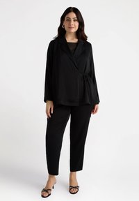 Jachetă neagră cu detaliu legat, asortată cu pantaloni negri. Material neted, mâneci lungi și sandale negre cu talpă ușoară și accente decorative.