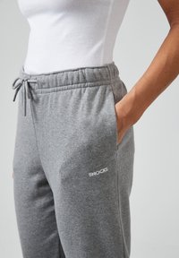 Graue Baumwoll-Sweatpants mit elastischem Bund, Kordelzug und Fronttaschen. Über dem Oberschenkel befindet sich ein weißes Logo.