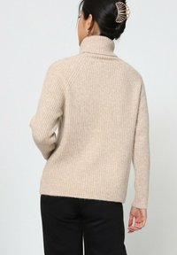 Pull beige à côtes avec un col montant, présentant des manches longues et une coupe décontractée. Texture douce et design minimaliste, porté avec un pantalon sombre.