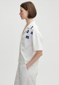 Blusa blanca de manga corta en tejido texturizado, con flores azules bordadas en los hombros, con un cierre frontal de botones y un corte relajado.