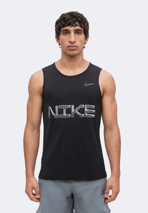 Mężczyzna w czarnym bezrękawnym podkoszulku Nike z dużym logo i szarych szortach, stoi zwrócony przodem na jednolitym tle.