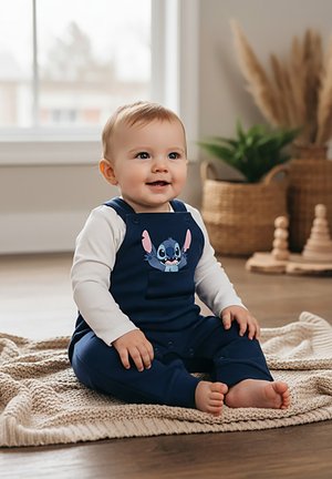 Bambino sorridente con salopette blu navy con un personaggio dei cartoni animati, seduto a piedi nudi su una coperta lavorata a maglia in una stanza luminosa e accogliente con piante.