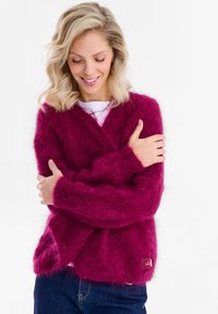 Kobieta o blond włosach, ubrana w różowy, puszysty kardigan, niebieskie dżinsy i białą koszulę, uśmiechająca się, z lekko skrzyżowanymi rękami na piersi.