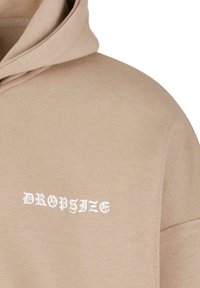 Sweat-shirt à capuche beige en tissu mélangé coton avec une texture douce. Présente un texte brodé blanc "DROPSIZE" sur la poitrine.