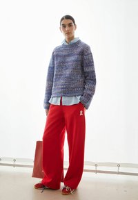 Gebreide trui in blauw- en paarstinten, gecombineerd met felrode, wijde broek met een geborduurd lint. De schoenen zijn rood met blauwe veters.