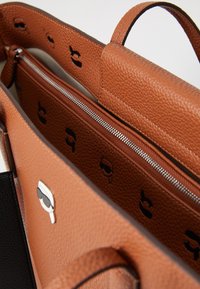 Sac à main en cuir marron texturé avec une poche zippée, présentant un intérieur doublé avec des motifs de logo noirs et des poignées supérieures doubles.