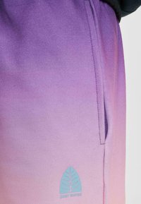 Pantaloni della tuta in tinta unita viola con una texture morbida; presentano una tasca laterale e un logo a foglia blu con il testo "JUST RHYME" all'orlo.