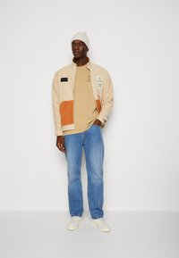 Tan fleece jacket with an orange patch pocket, beige t-shirt, blue jeans, and white sneakers, topped with a light knit beanie.