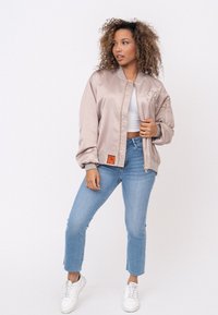 Giacca bomber di raso rosa chiaro con polsini grigi e tasca laterale con zip, caratterizzata da un'etichetta arancione e un logo sul petto. Indossata con jeans blu.