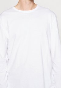 Camiseta blanca de manga larga con cuello redondo, hecha de material de algodón suave, con una textura lisa y sin patrones ni acentos visibles.