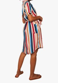 Donna che indossa un vestito a righe verticali multicolori con maniche corte, una cintura in vita e tasche laterali, abbinato a sandali marroni.