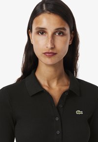 Svart ribbad pikétröja med krage, med en grön broderad krokodil-logotyp och fem svarta knappar framtill.