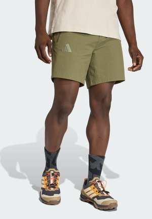 Männliches Model trägt olivgrüne Sportshorts, schwarze gemusterte Socken und beige-orange Laufschuhe, steht vor einem einfarbigen Hintergrund.