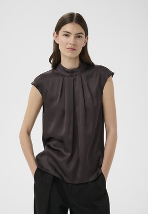 Vrouw met stijlbruin haar draagt een mouwloze donkere zijden blouse met hoge kraag en losse zwarte broek, staand met handen in de zakken.