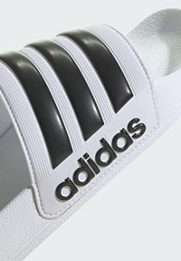 Hvit slidesandal med tre svarte striper, strukturert rem og glansfull logo. Laget av syntetisk materiale, med en formet fotseng.