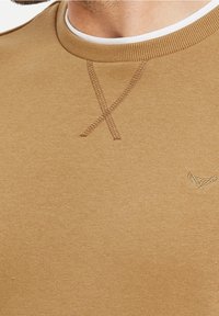 Sudadera marrón con cuello redondo, patrón de costura cruzada sutil y un pequeño logotipo en relieve en la parte inferior derecha. Textura suave.