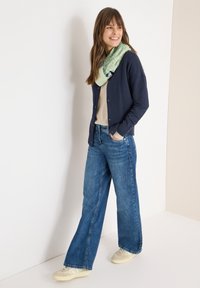 Marineblauw vest over een beige top, gecombineerd met blauwe wijde jeans en een groene sjaal met patroon. Casual sneakers maken de outfit compleet.