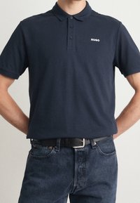 Granatowa koszulka polo, materiał bawełniany, trzy guziki z przodu, ściągacz przy kołnierzyku, biały logo "Hugo" na lewej piersi, zestawiona z ciemnoniebieskimi dżinsami.