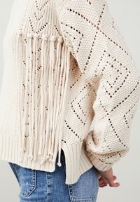 Maglione beige lavorato a maglia con un motivo a trama aperta e design geometrici, con nappine laterali e polsini e orlo a coste.