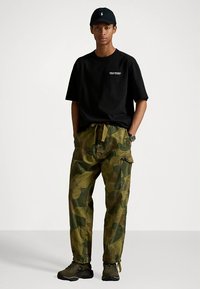 Μαύρο κοντομάνικο μπλουζάκι με λογότυπο, πράσινο καμουφλάζ παντελόνι cargo με τσέπες και σκούρα αθλητικά παπούτσια, φορεμένα με μαύρο καπέλο.